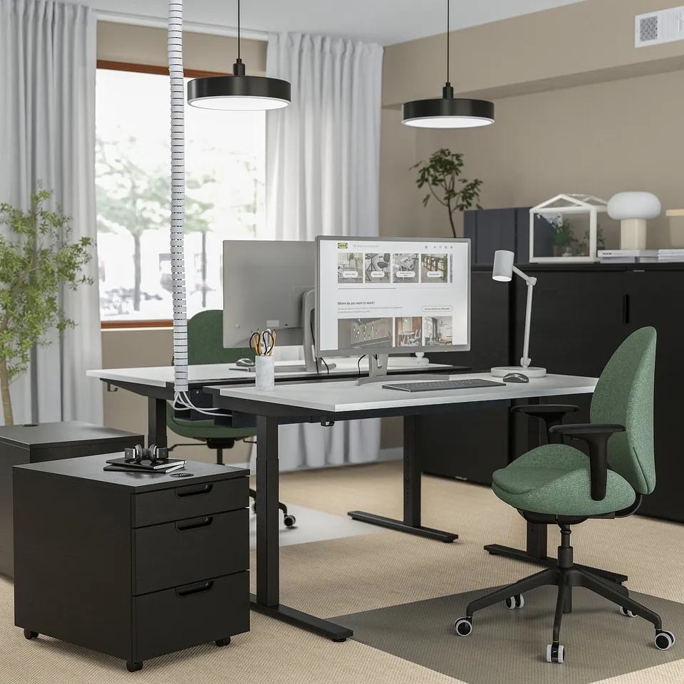 MITTZON black height-adjustable office desk with green chair, dual monitors, and pendant lights. Sleek, modern design, larger than adjacent furniture.