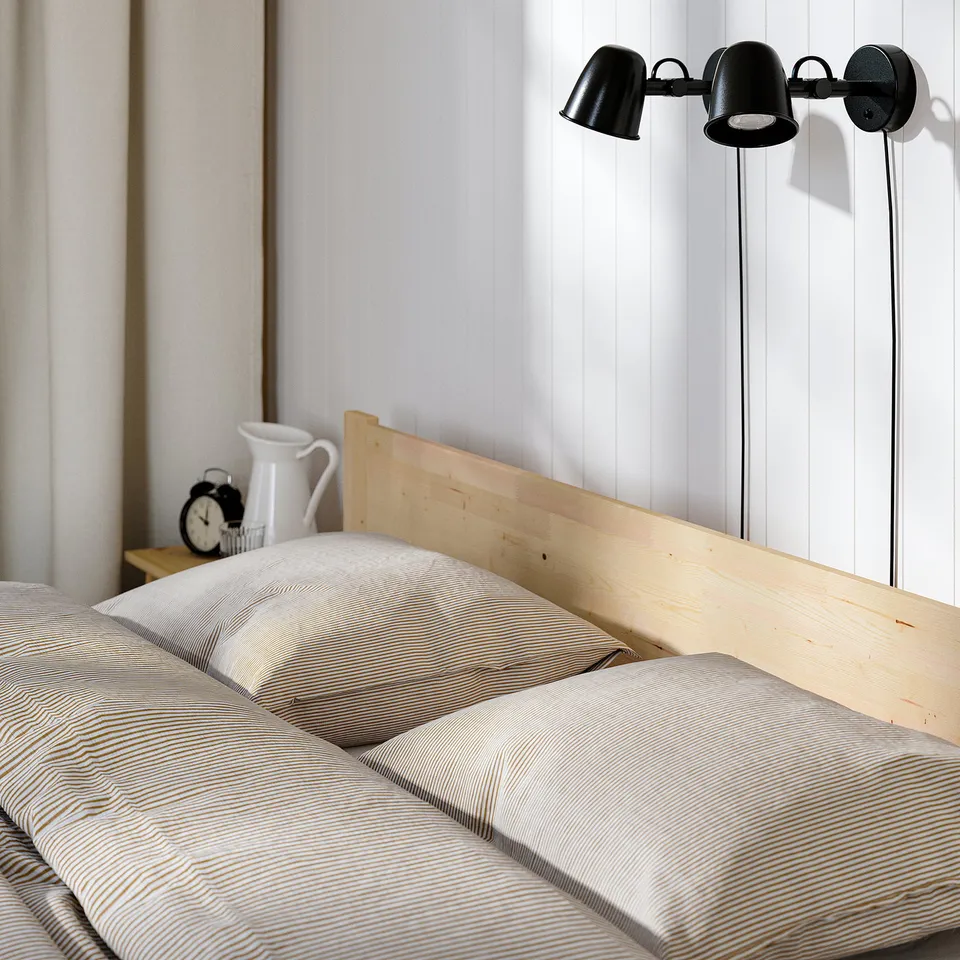 Minimalist bedroom with TAXÅSKLINT bed, white striped bedding, alarm clock, jug, and black wall lamps.