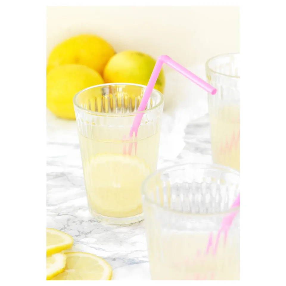 Two glasses with lemon juice and pink straws are placed on a marble table, surrounded by lemons.