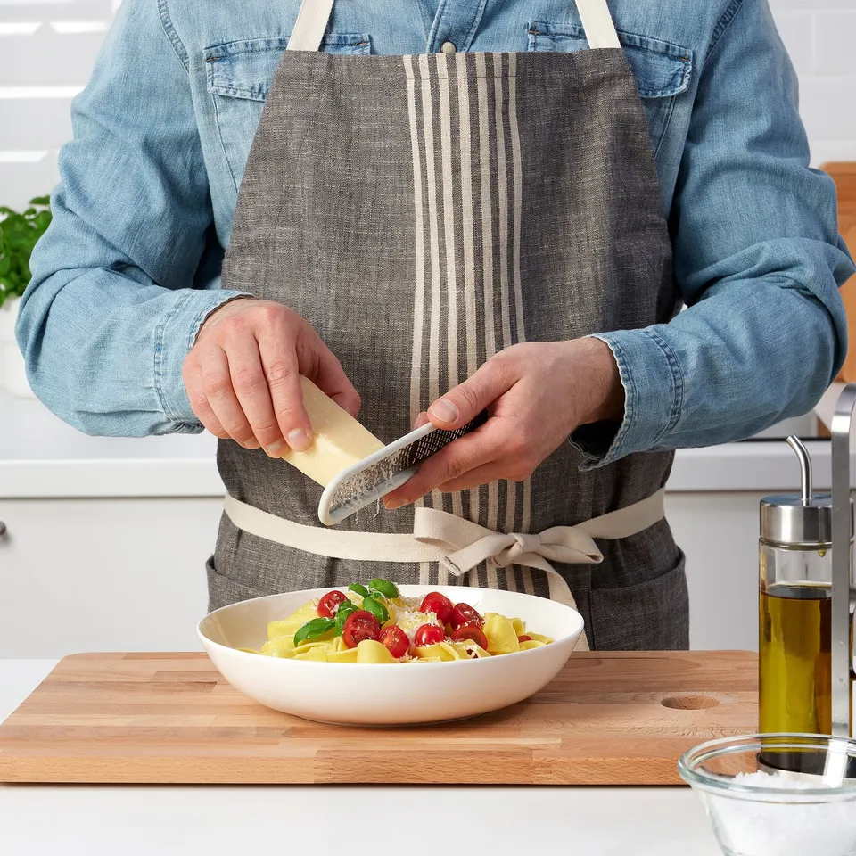 A person grates cheese over pasta with a small handheld grater on a kitchen counter.