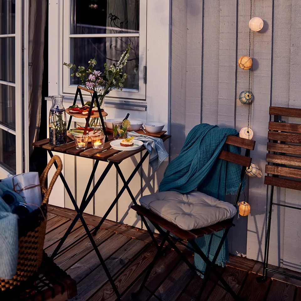 Small outdoor table set with string lights, a teal blanket, and wooden chairs beside a light blue wooden house.
