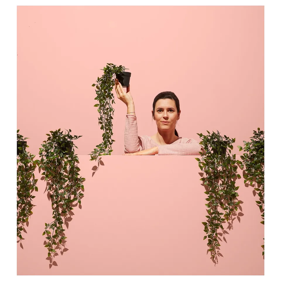 A person holding a green FEJKA artificial plant, set against a pink background with more green FEJKA plants displaying bright natural colours.