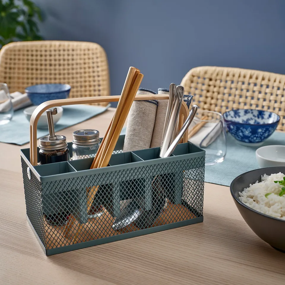 A VIMPELFISK green metal cutlery caddy with bamboo handle, holding cutlery and napkins, placed on a wooden table with a bowl of rice.