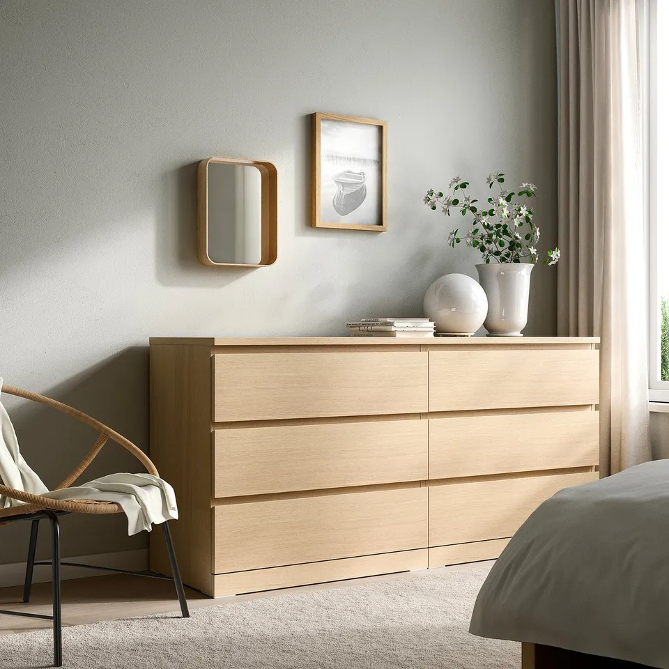 Minimalist bedroom with oak furniture: mirror, dresser, vase, chair, window.