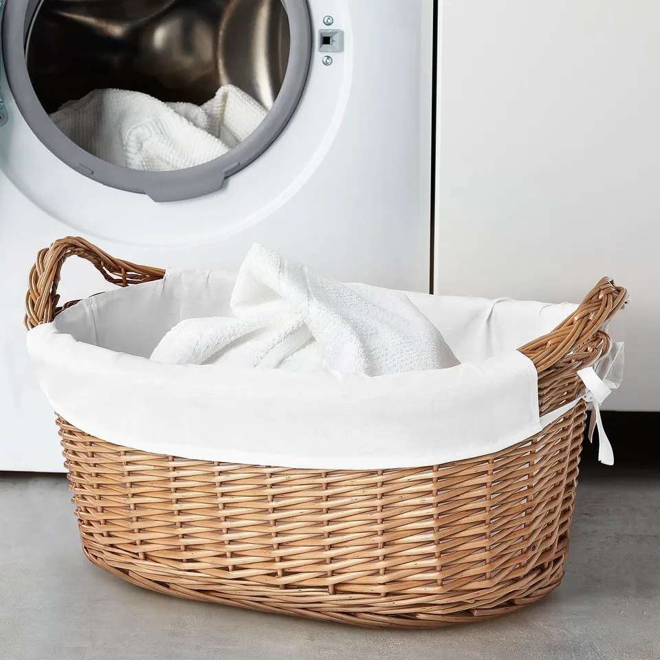 The TOLKNING laundry basket in natural willow weave with a large white inner bag. Circular shape, with sturdy handles, fits beside a washing machine.