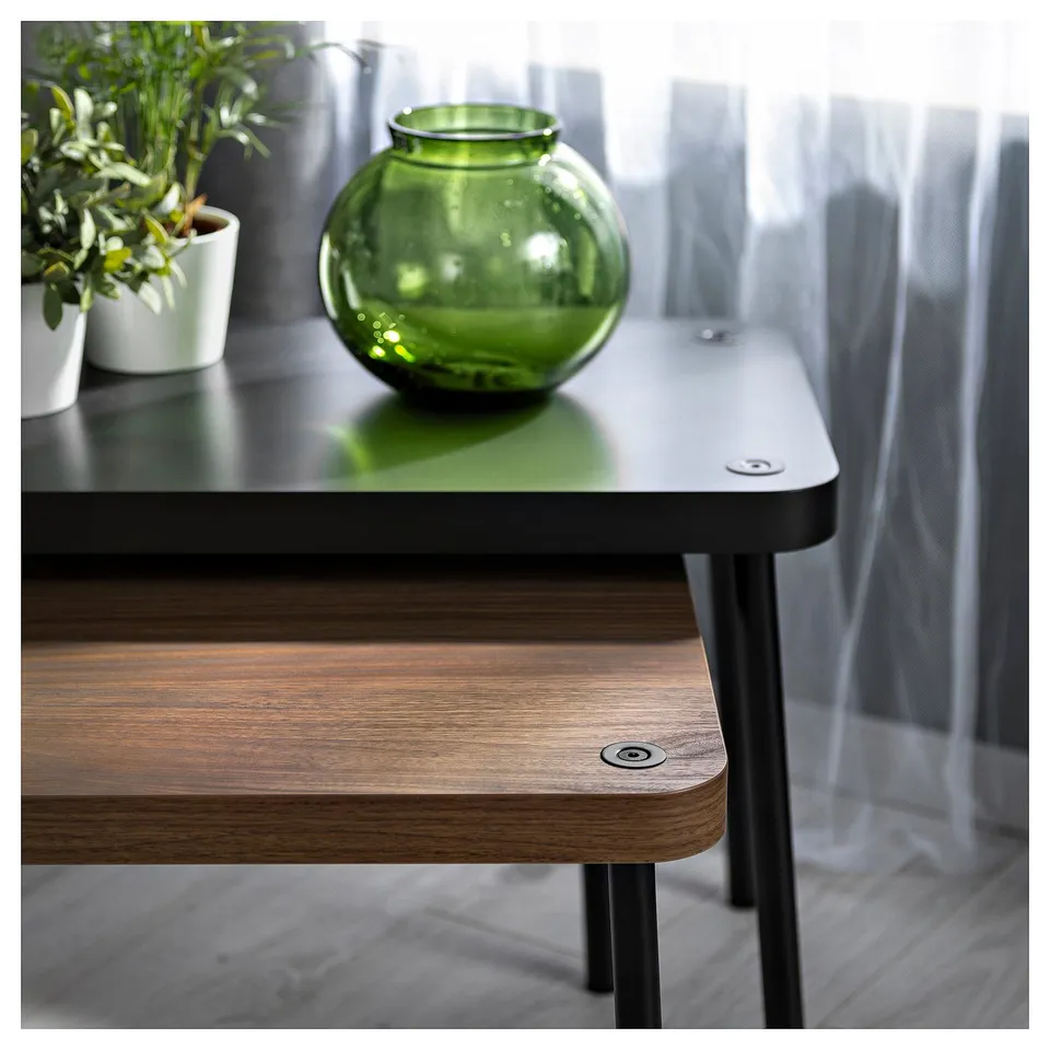 Two modern wooden nest tables with black metal legs. On top, a green vase and a potted plant. Tables have sleek, rounded edges.