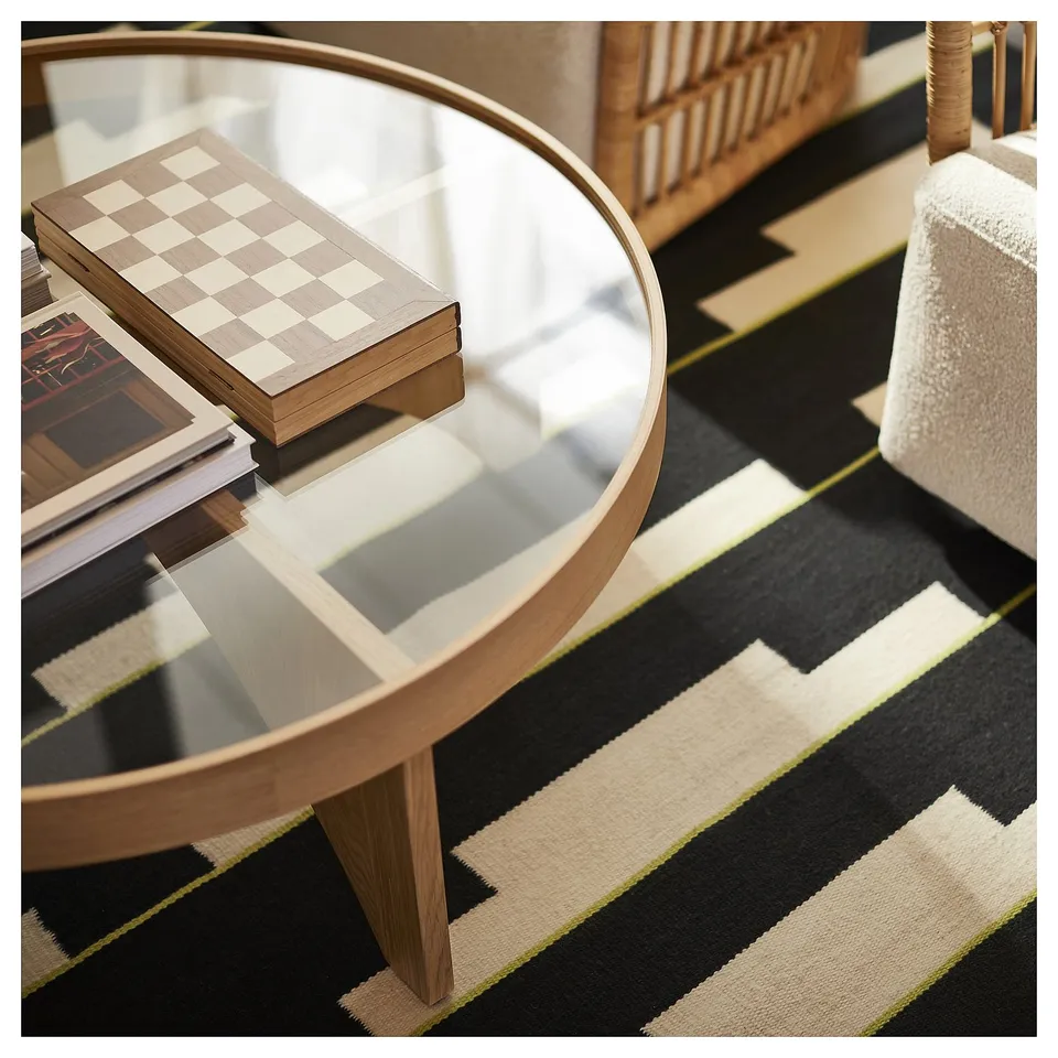 Modern living room with glass-topped table, chess set, books, geometric rug, wicker chair.