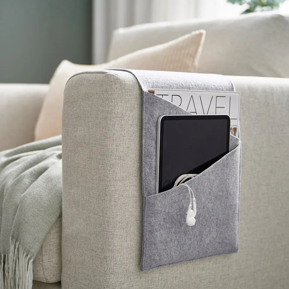 Light grey SILÄNG pocket organiser hangs on sofa arm, holding tablet, magazine, and earphones.