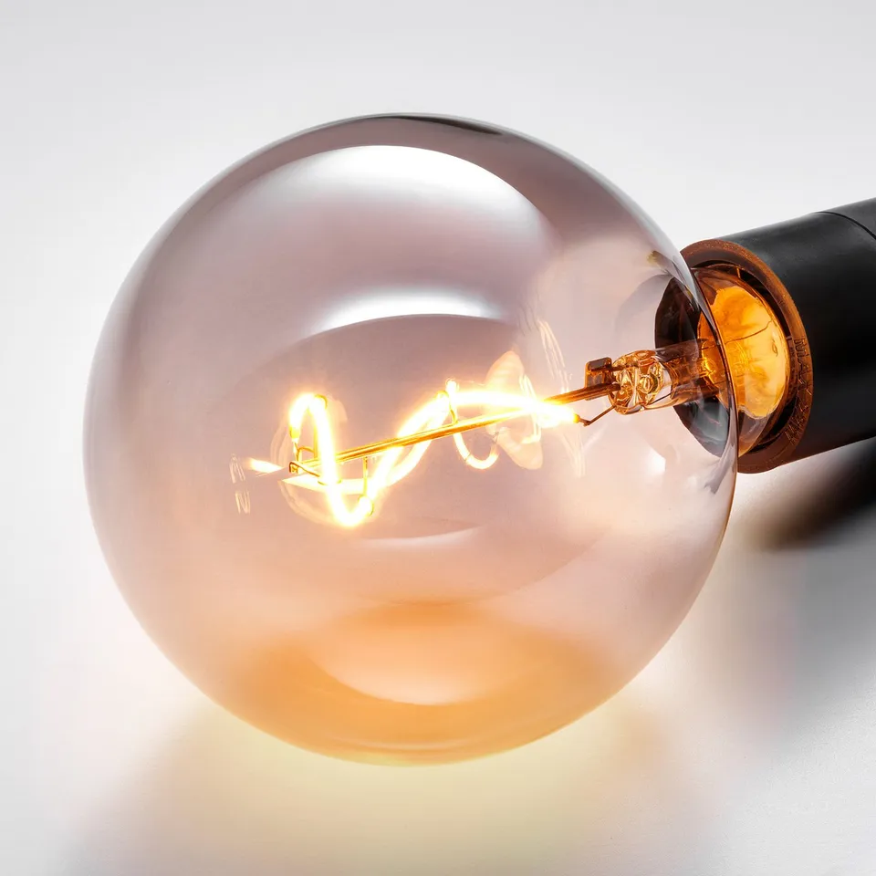 Close-up of a frosted glass bulb with a glowing spiral filament, emitting warm light. A black socket holds the bulb, suggesting a modern decor.