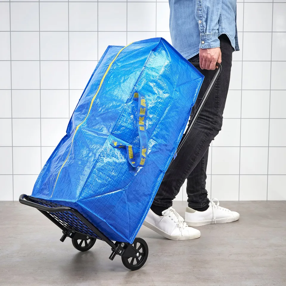 Person pulls large blue frakta bag on wheeled cart against tiled wall.