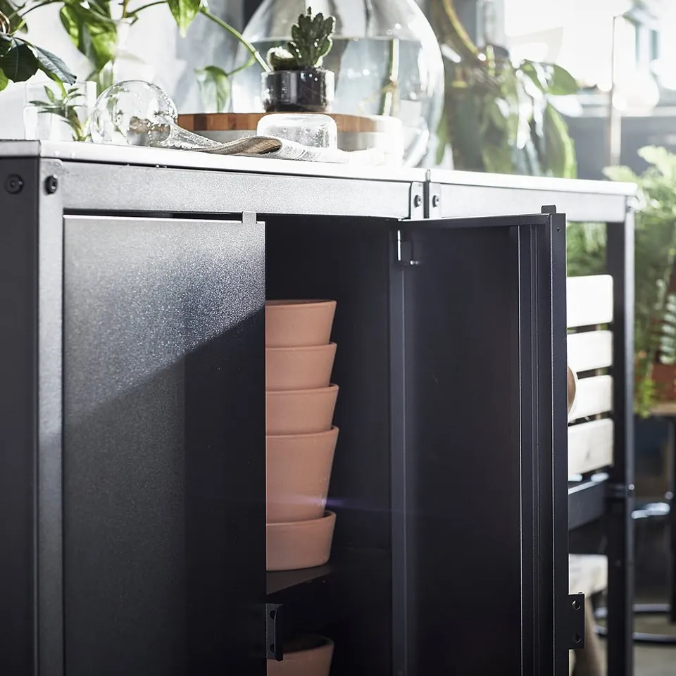 Black outdoor kitchen island, stainless steel top, stack of terracotta pots, decorative glass items, surrounded by plants.