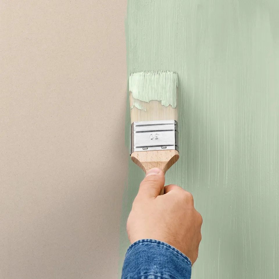 Person painting a wall using a paintbrush. The paint is being applied to the wall in vertical lines. The paint colour is a muted green shade.