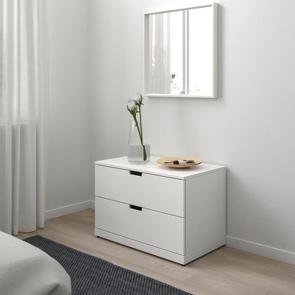 Minimalist bedroom with white NORDLI chest, mirror, and flowers.