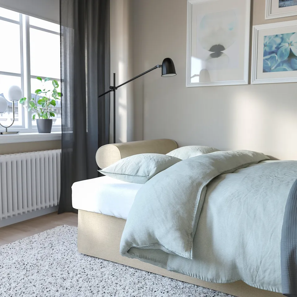 Modern bedroom with grey curtains, bed made with light blue blanket, green pillow, art on wall, plant by window, white radiator, lamp near bed.
