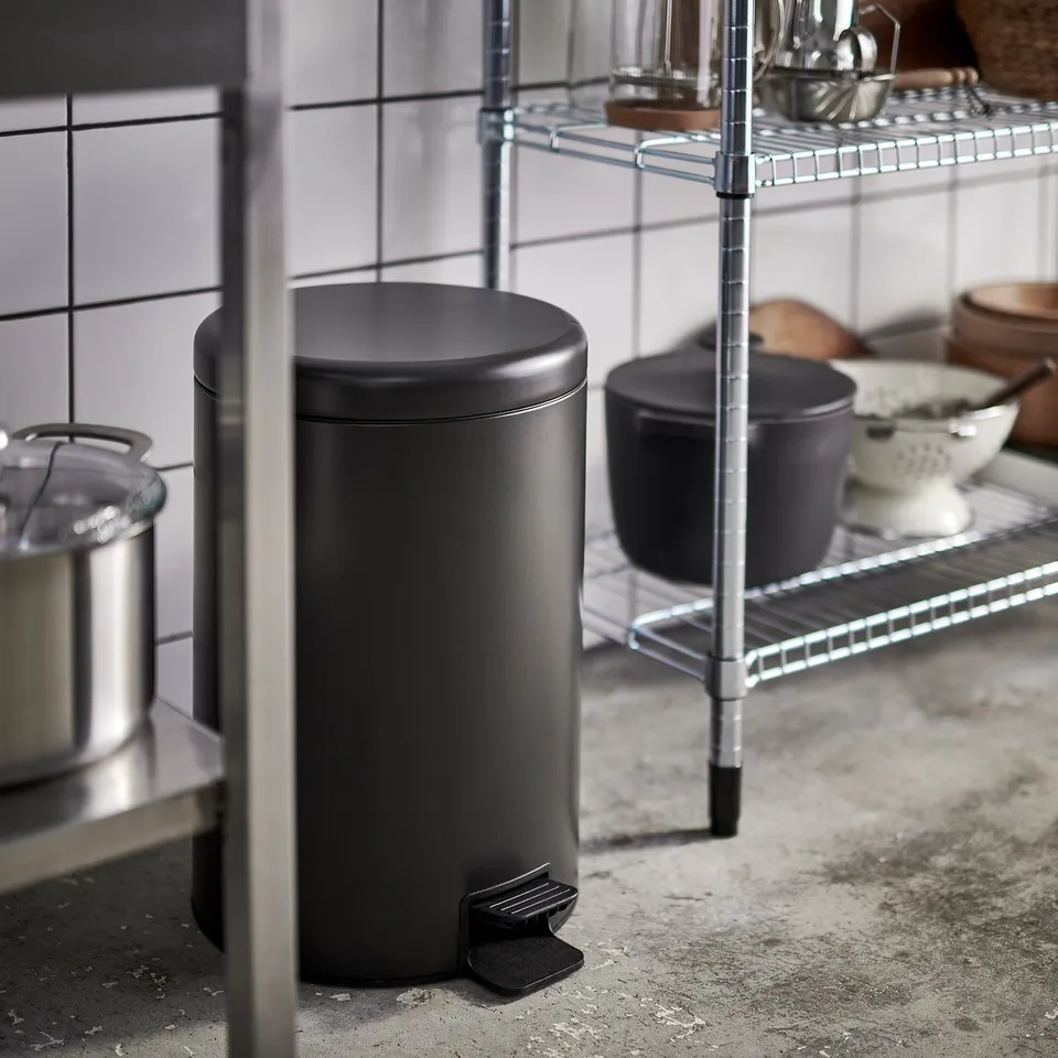 Small black pedal bin in kitchen next to wire rack with cans and bowls.