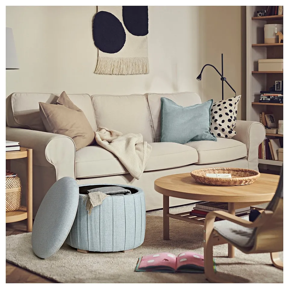 Herringbone-patterned beige sofa with blue and polka-dotted pillows, light blue round storage ottoman, wooden coffee table with tray, rug, and books.