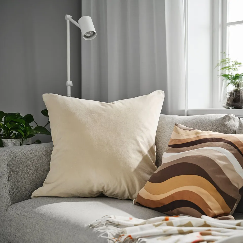 A large SANELA beige cushion cover on a light grey couch, surrounded by other cushions and a blanket, with plants and a lamp nearby.