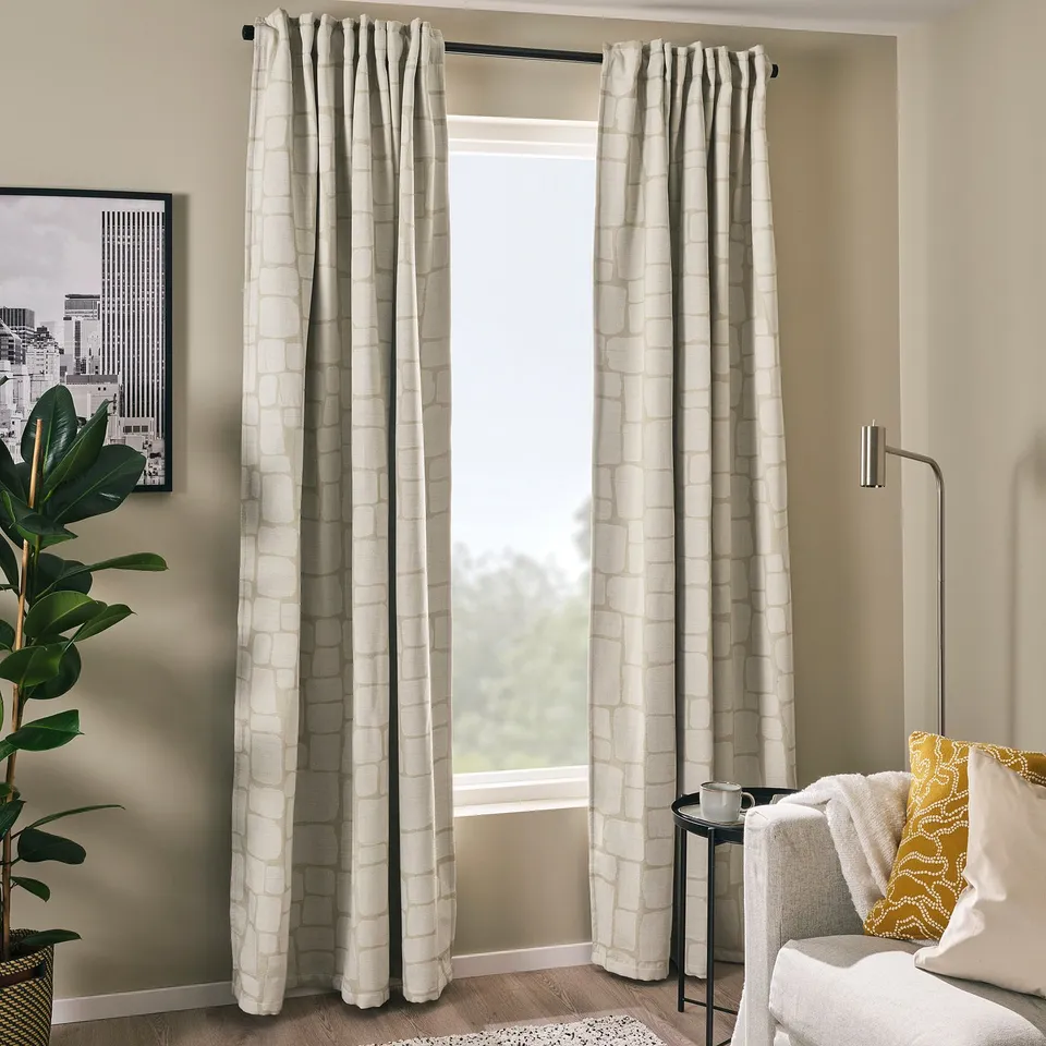 Beige LÖNNSTÄVMAL block-out curtains with jacquard pattern between window and wall plant, partially covering white sofa with yellow pillow.