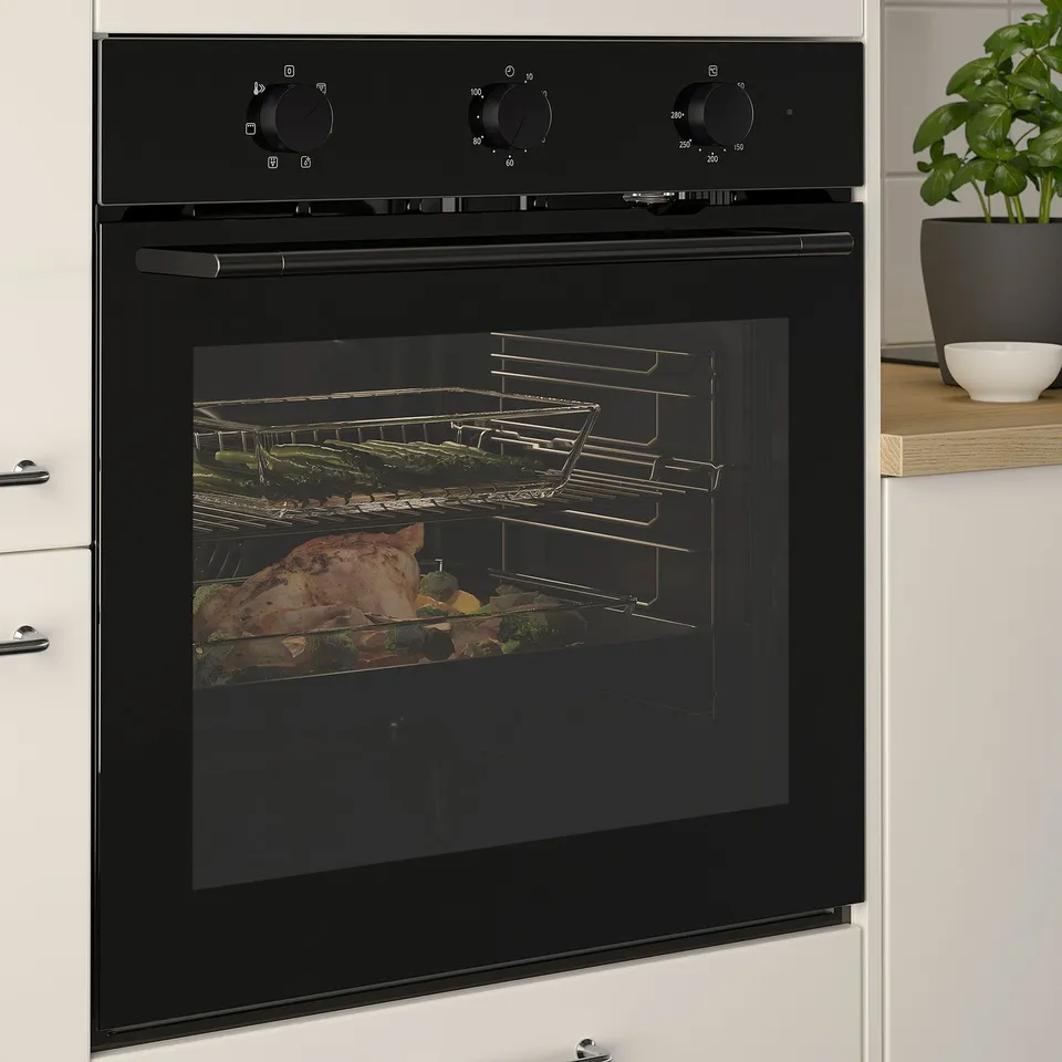 Black oven with smoked glass door, cooking multiple dishes on racks, placed in white kitchen cabinetry.