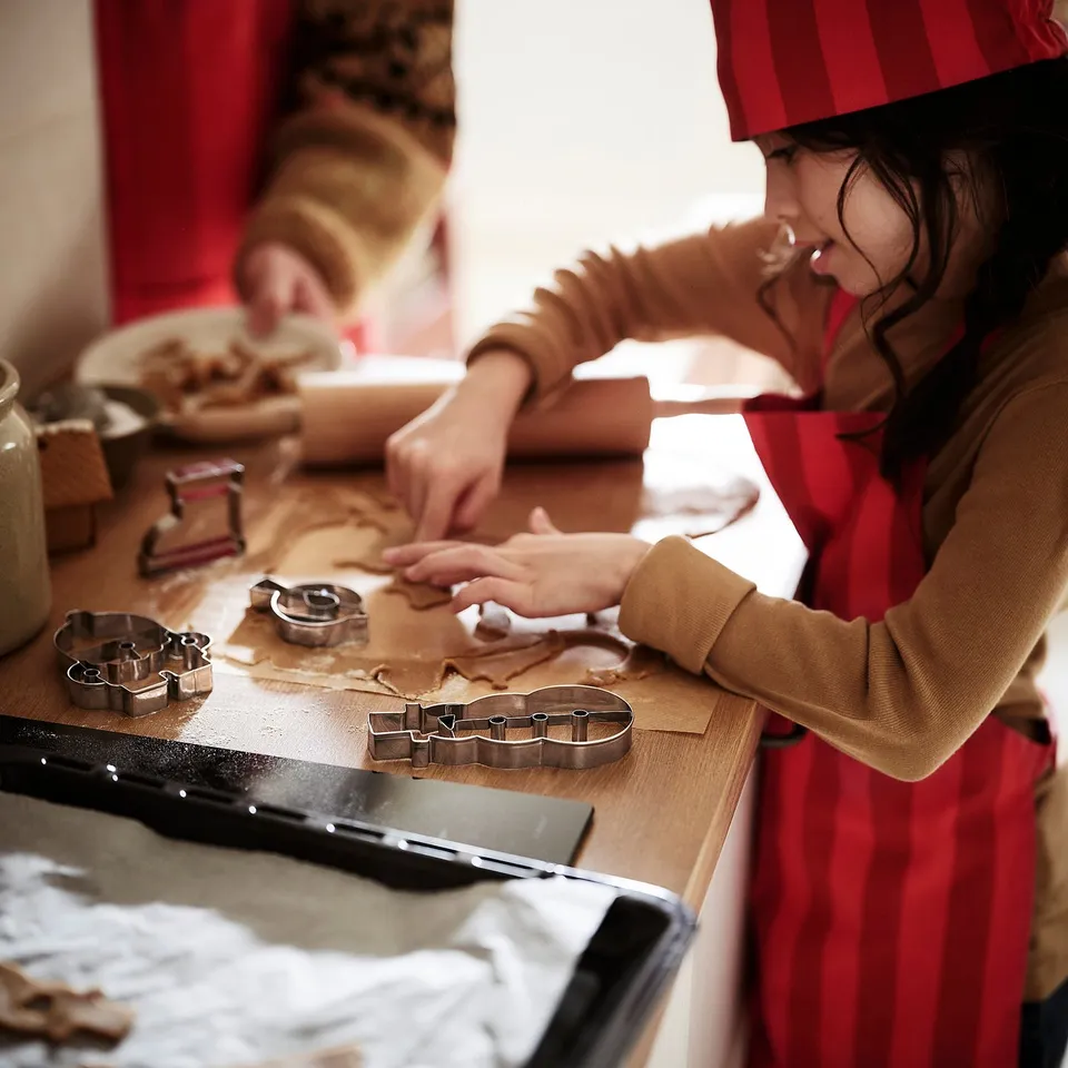 Child wearing red, striped chefs hat bakes cookies using cutters. The hat fits various ages, enhancing holiday baking fun.