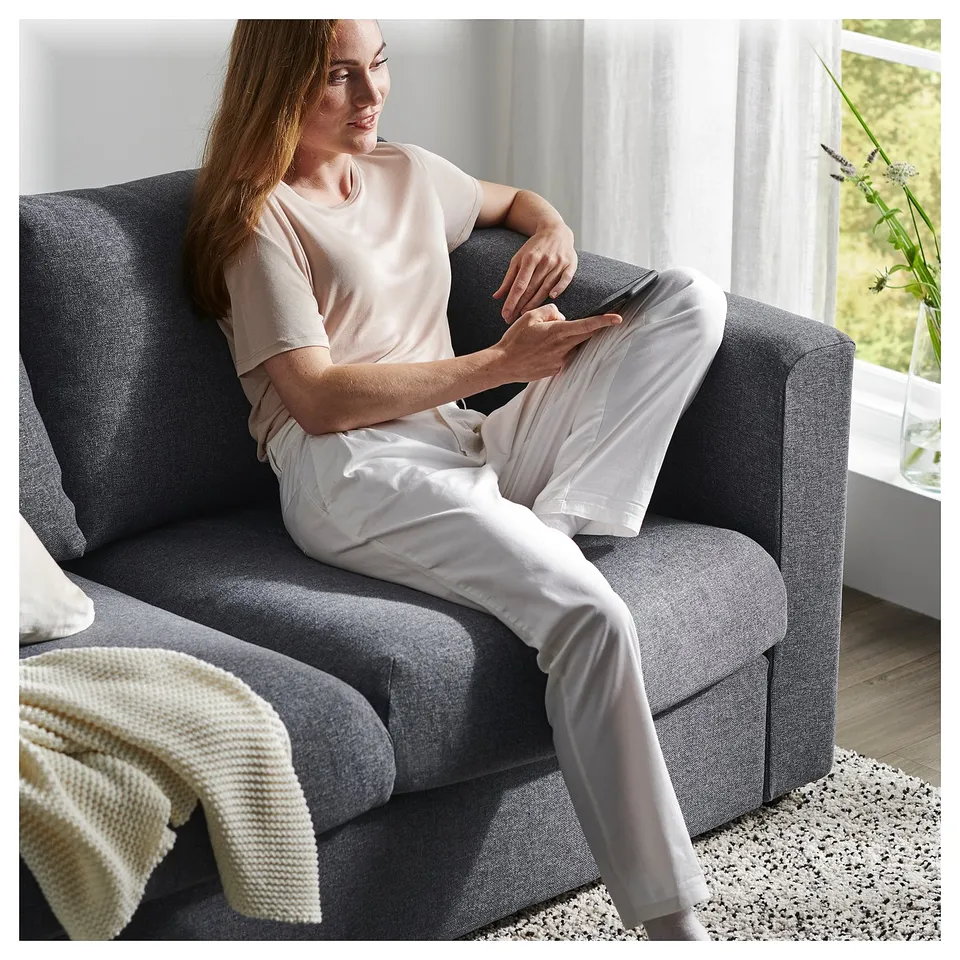 A person relaxes on a grey VIMLE sofa with a white throw pillow, holding a phone, wearing a beige shirt and white pants.