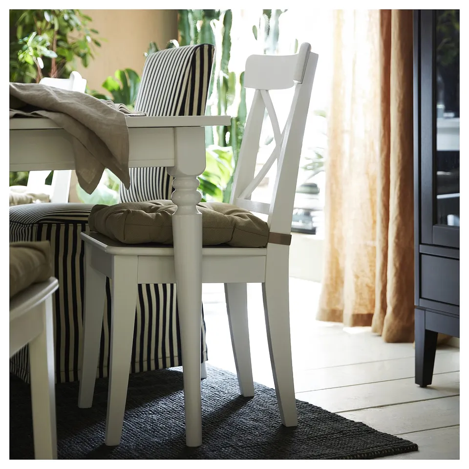 Clean dining area with white chairs, stripped cushions, green plants, and beige curtains on bright windows. Cosy natural rug on floor.