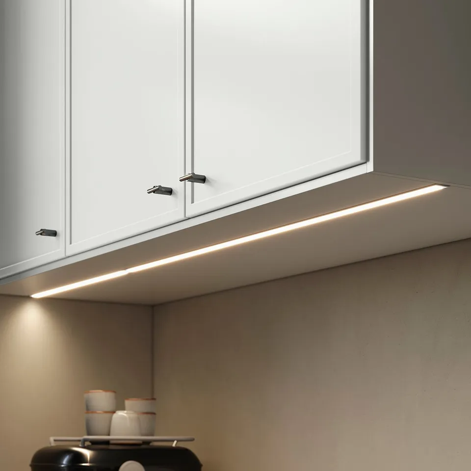 Modern kitchen with white cabinets, lighting, minimalistic handles, counter with mugs and coffee maker.