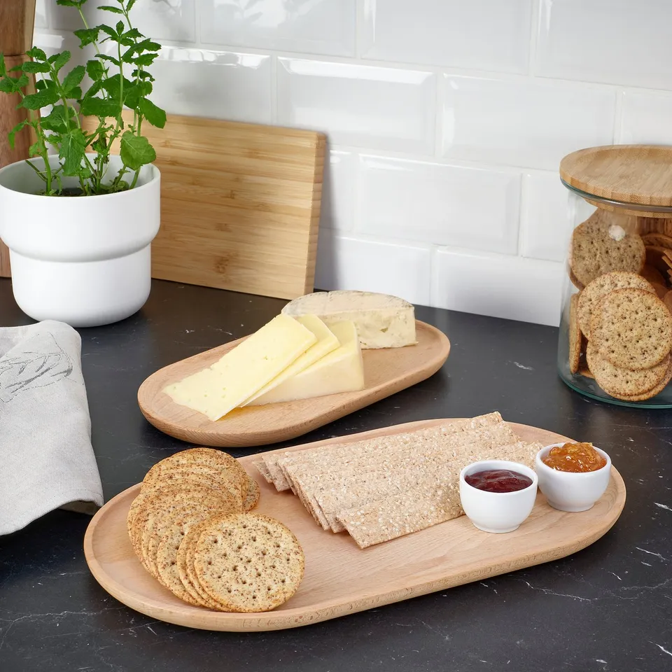 Two wooden trays hold cheese, crackers, and dips. A plant and towels are nearby on a dark countertop.