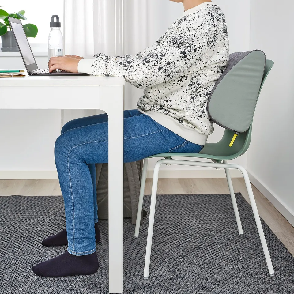 Person sitting at desk, using ÖVNING footrest. Footrest supports feet, helping maintain good posture. Desk has laptop, water bottle, and plant.
