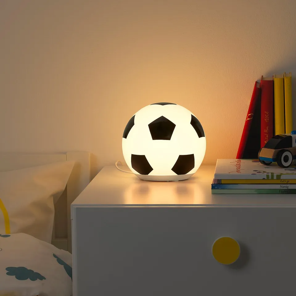 Football-shaped ÄNGARNA lamp on bedside table with books and toy, glowing softly.