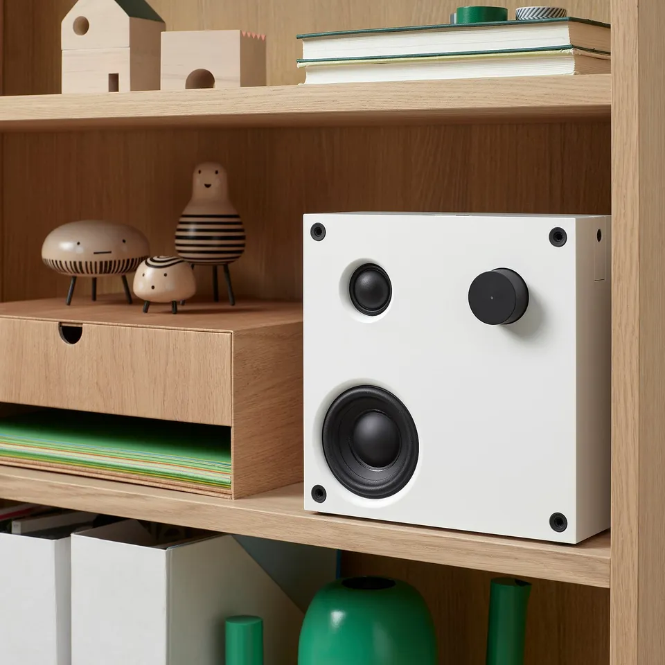 A white VAPPEBY bluetooth speaker on a wooden shelf, between ceramic figurines and vinyl records.