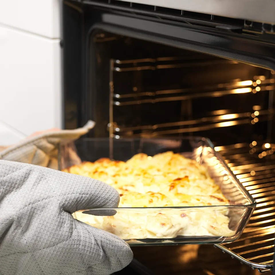 Gloved hand removing baked dish from oven. Golden, bubbly top suggesting a casserole. Oven rack visible, indicating baking process.