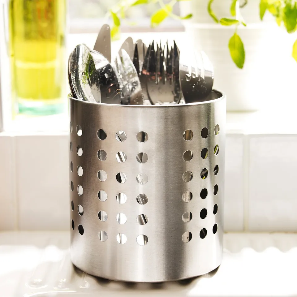 Metal cutlery holder with holes, placed on counter storing utensils.