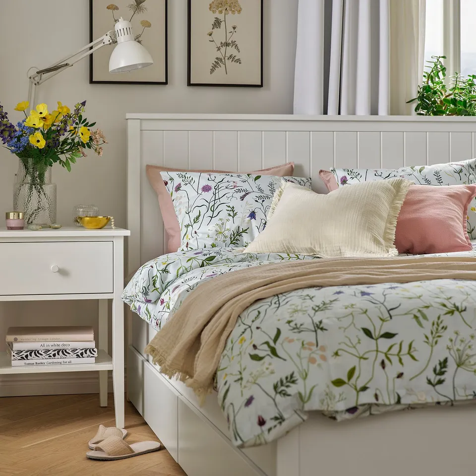 Cosy bedroom with MATTOXBÄR blue floral bedding, white pillows, pink accents, and a bedside table with lamp, flowers, and books.