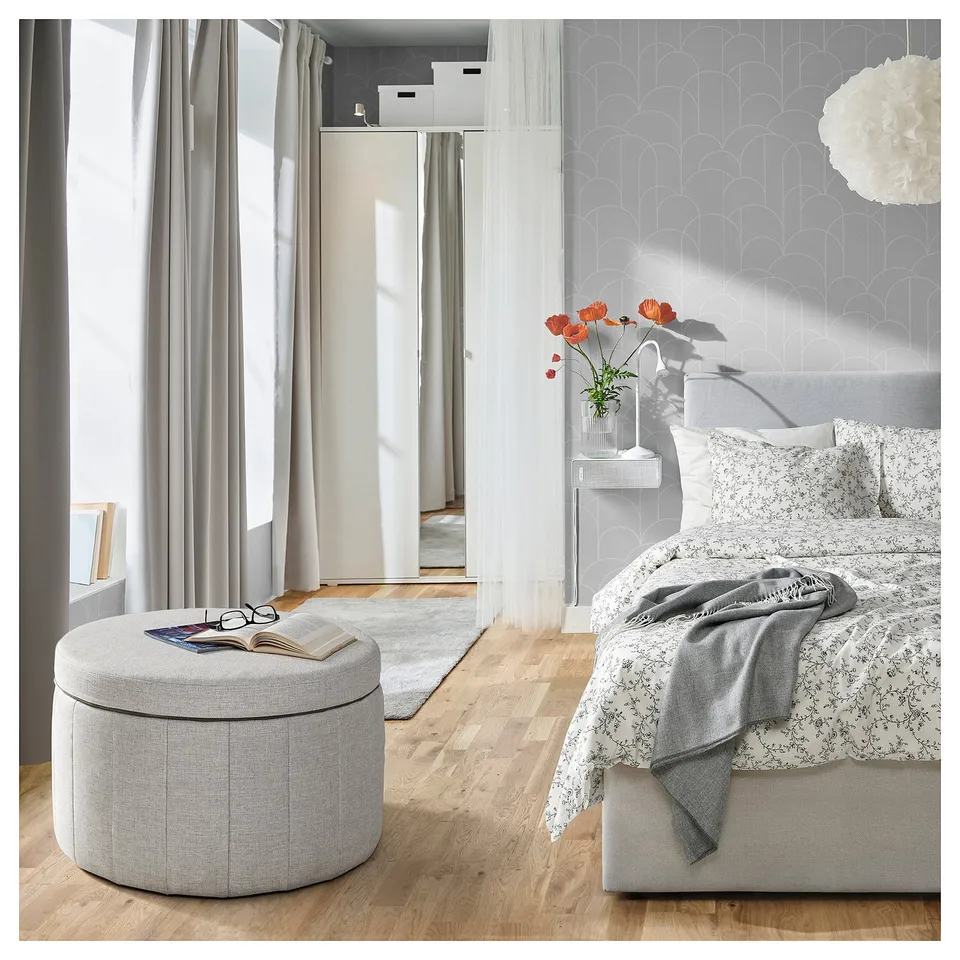 Modern bedroom: light wood floor, white bed, grey throw, grey ottoman, books, red flowers on bedding, white wallpaper, pendant light.