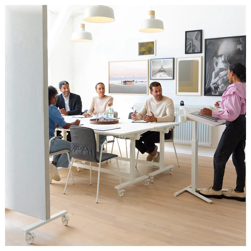 Meeting around MITTZON table in minimalist room with lamps and art. Person uses laptop separately.
