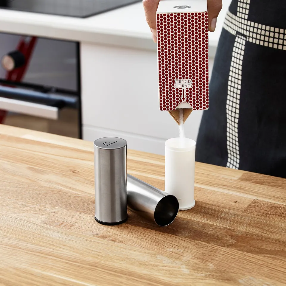 Person pouring contents from a red box with dots into a white container on a wooden table, assisted by metallic and black accessories.