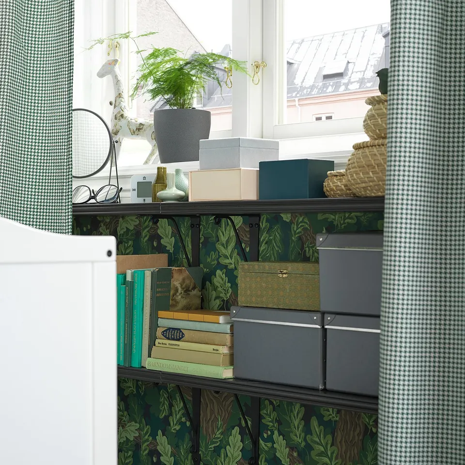 Bookshelf with green leafy wallpaper, books, vases, plant, boxes; white window frames.