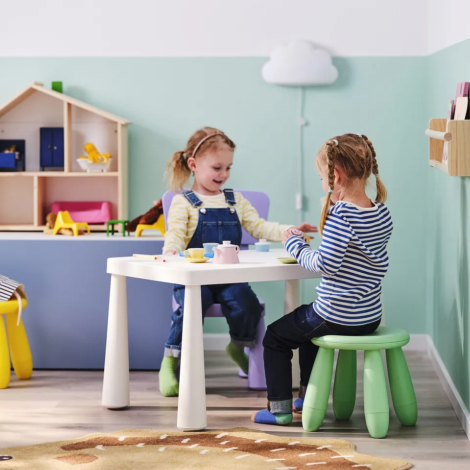 Two girls play with teacups at a white table in a colourful playroom.