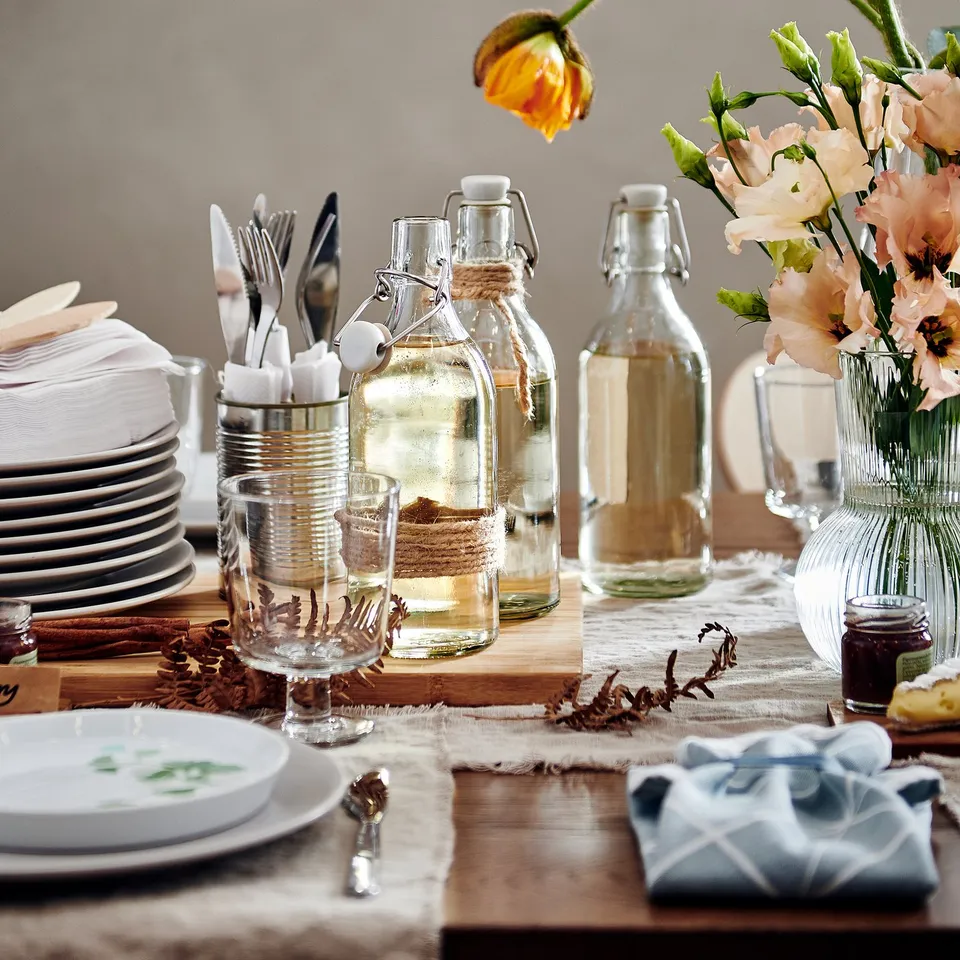 Table set with plates, cutlery in glass jars, floral centrepiece in KORKEN bottles, jam, and napkins.