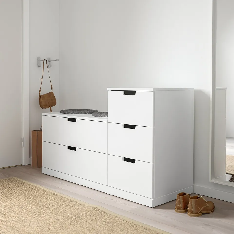 White NORDLI 3-drawer chest in minimalist room. Black handles, between wall & door, near coat rack & rug.