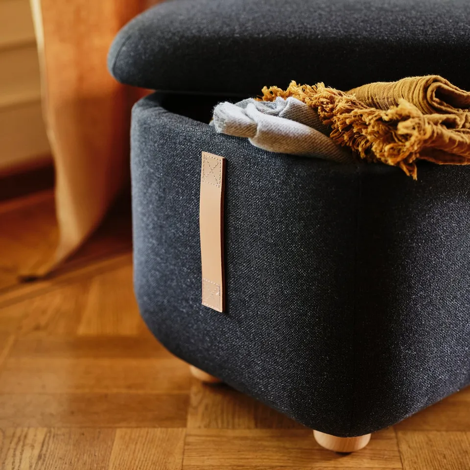 Dark blue fabric-covered footstool with wooden legs, slightly open to reveal folded textiles inside. Label on side shows a recycling logo.