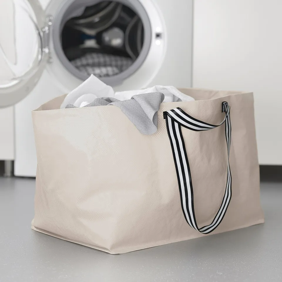 White GÖRSNYGG laundry bag near washing machine. Rectangular, durable, striped handles.