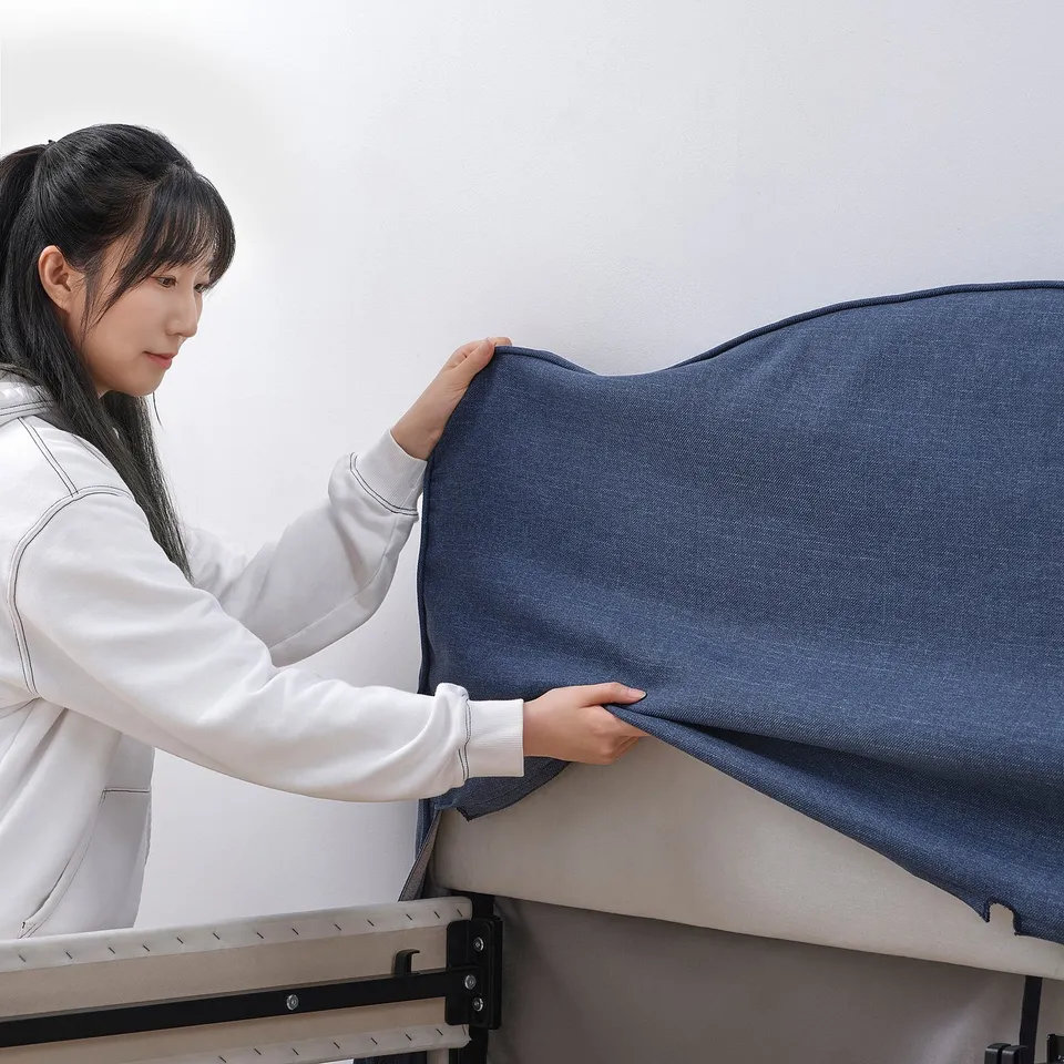 Person adjusting grey upholstered bed with removable cover against wall.