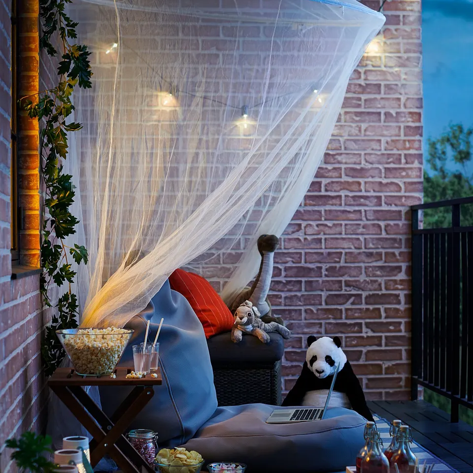 Cosy patio setup with black and blue cushioned chair, white net, string lights, snacks, drinks, and stuffed animals for relaxation.