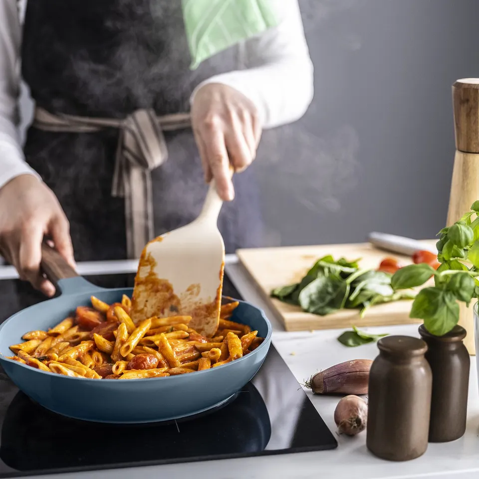 Person cooking pasta in a grey-blue frying pan with wooden handles. Ingredients and utensils are on the side.