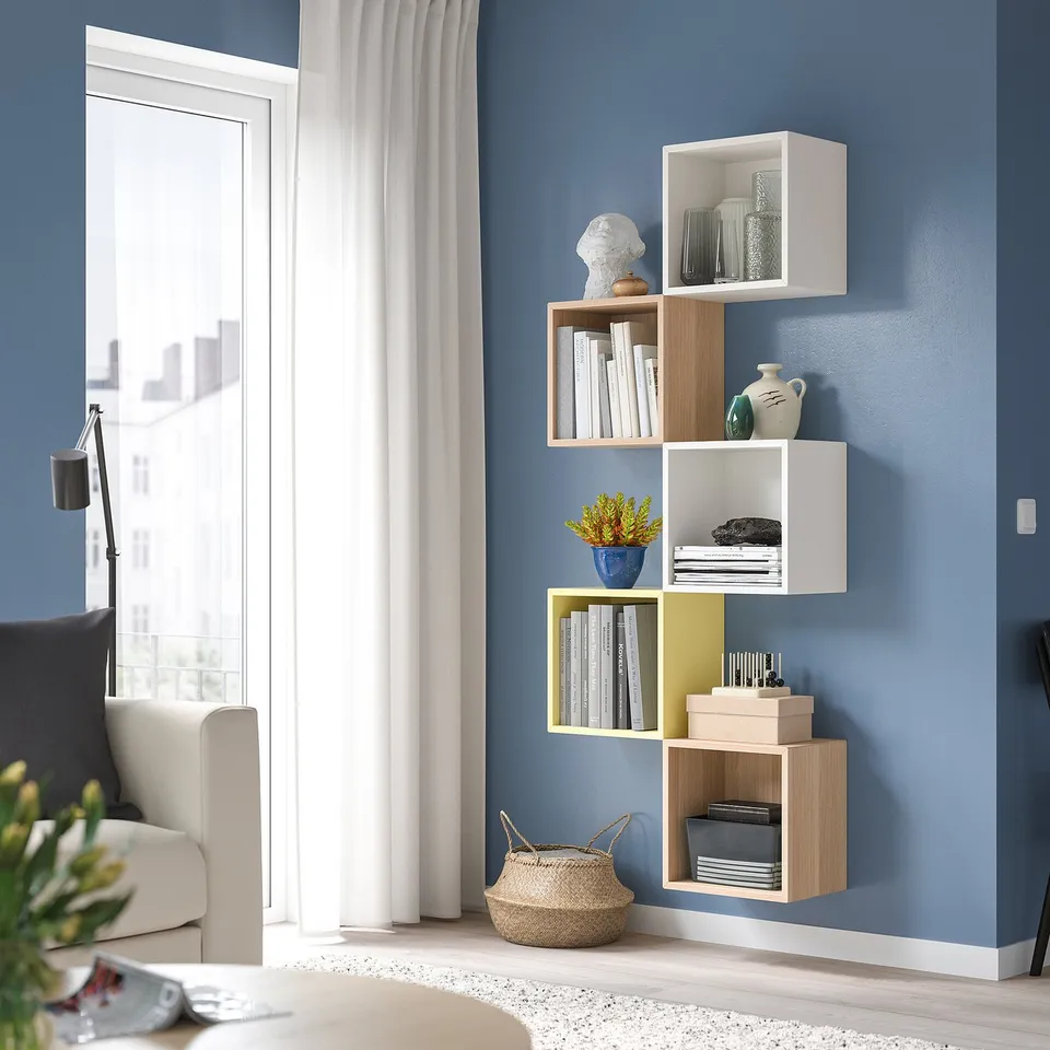 Modular shelves with books, vases, plants against blue wall. Clean, rectangular, light wood, mustard, window light.