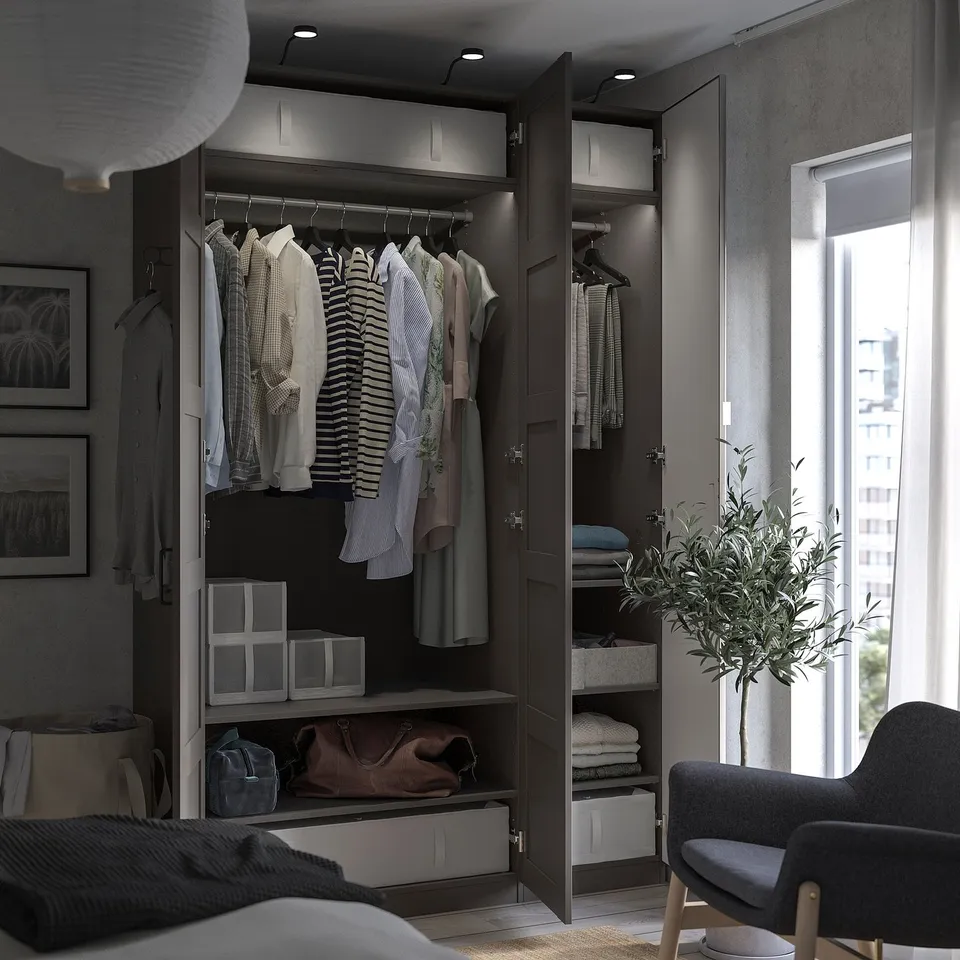 Modern bedroom with open black PAX wardrobe displaying clothes, accessories, and lit shelves. Large window, grey chair, and potted plant visible.