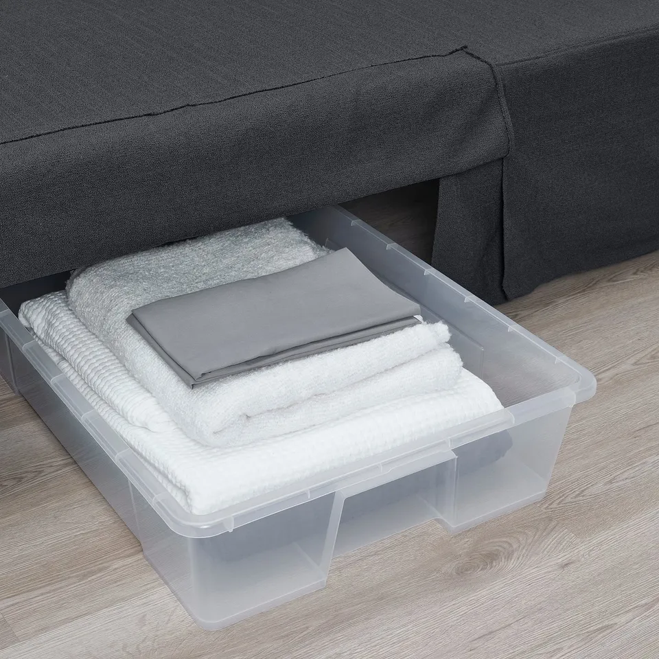 Clear plastic storage bin under grey sofa holds neatly folded white and grey towels.