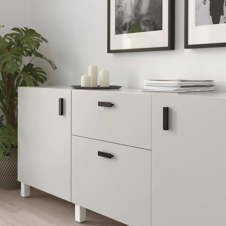 White cabinet with black handles, holding candles and magazines, next to a plant.